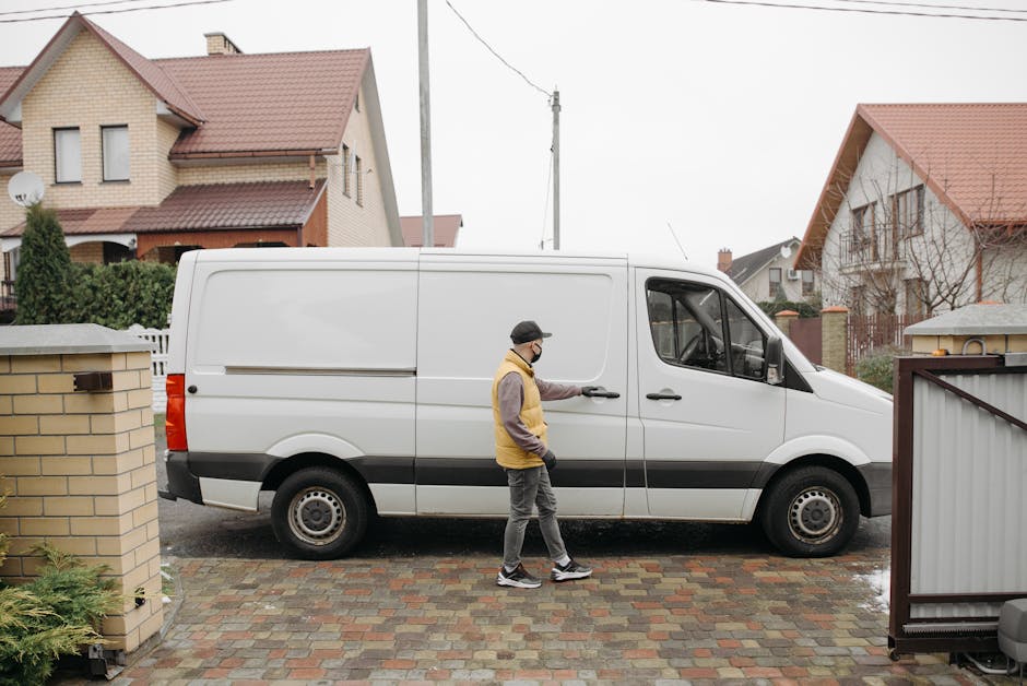 A man wearing a grey and yellow jacket, grey pants, and a black cap is standing outside a residential property, opening the sliding door of a white panel van parked on a paved driveway. The van is positioned in front of a brick wall and a metal gate, with the van's side door partly open, indicating the loading process. In the background, there are several detached houses with pitched roofs, some with visible windows and dormers, and an overcast sky. The scene takes place in a suburban neighbourhood, with residential buildings and utility poles visible. The man appears to be preparing for furniture transport or a home relocation, with the van ready for moving items, and the environment prepared for the logistics involved in packing and moving household goods. Man and Van Bexleyheath’s service relates to such removals, supporting efficient and safe furniture transport.