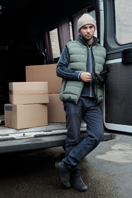 A man with a light beard and neutral expression standing at the rear of a black van, which is parked outdoors on a concrete surface. He is wearing a grey knitted beanie, a green padded vest over a navy blue jacket, dark trousers, and black work boots. The van's rear doors are open, revealing several cardboard boxes of various sizes stacked inside, some sealed with packing tape. The boxes are positioned on the van's loading area, which is partly visible, with a few larger boxes resting against the van's interior wall. The scene appears to be part of a home relocation or furniture transport process, with the man preparing to load or unload items as part of a professional removal service. The environment is well-lit, with natural daylight and a neutral background, aligning with house removals and packing activities typically managed by companies like Man and Van Bexleyheath.