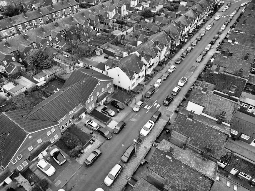 A narrow residential street with closely spaced brick terraced houses on the left side, featuring small front gardens enclosed by black metal fences and white-framed windows. On the right side, there