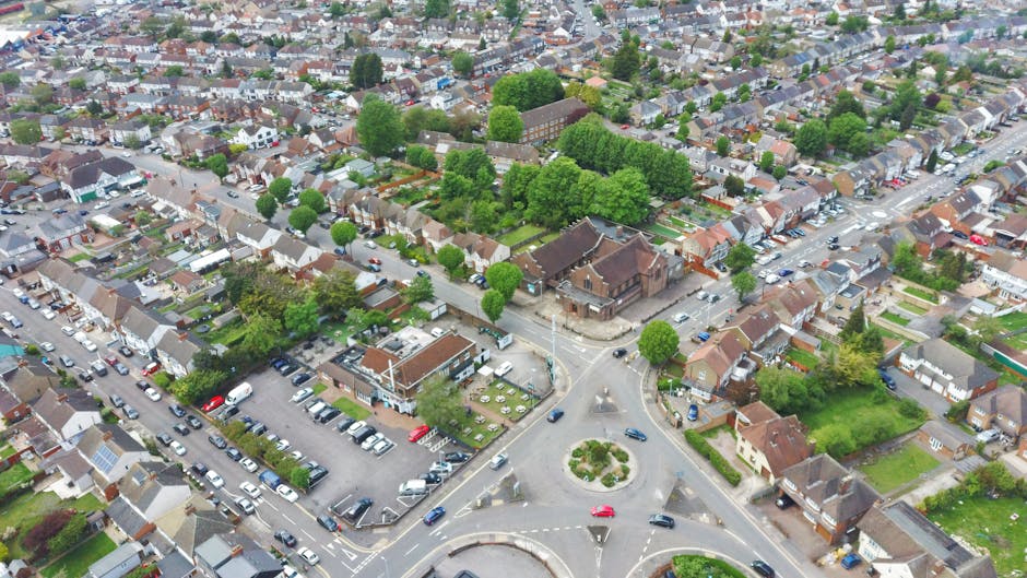 An aerial view of a residential neighbourhood featuring a roundabout at its center, surrounded by a mix of terraced and semi-detached houses with tiled roofs and small gardens. Several streets radiate from the roundabout, lined with parked cars and green trees, including some larger, leafy trees creating a canopy over the roads. In the foreground, there is a parking area filled with various vehicles near a cluster of buildings, possibly indicating a commercial or communal space. The scene is well-lit with natural daylight, showcasing the orderly layout of the streets and the lush greenery. This image captures the typical environment where house removals and furniture transport may occur, with visible access points for moving vehicles to facilitate home relocation activities. Man and Van Bexleyheath often coordinates with such settings for efficient packing, loading, and transportation of household items during moves.