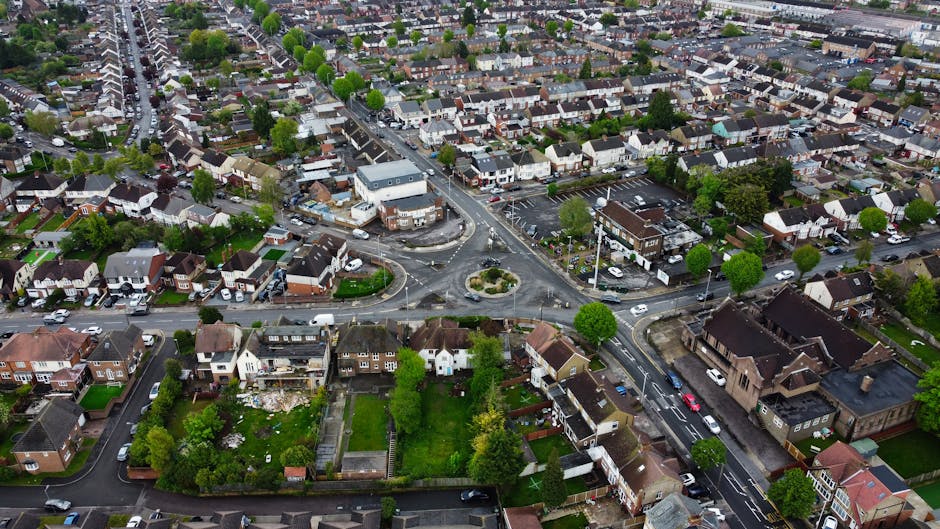 An aerial view of a suburban neighbourhood showing a mix of detached, semi-detached, and terraced houses with front gardens, tree-lined streets, and parked cars. The image captures a roundabout at the intersection of several roads, with some vehicles navigating the traffic flow. There are visible parking areas, and the houses have varied roofing materials, including slate and tiled roofs. The scene appears to be taken during daylight with natural lighting, highlighting the green foliage and well-maintained properties. In the context of home relocation or furniture transport, this image exemplifies the typical environment where professional removals, such as those offered by Man and Van Bexleyheath, coordinate loading and unloading goods, packing materials, and furniture within the residential suburban setting.