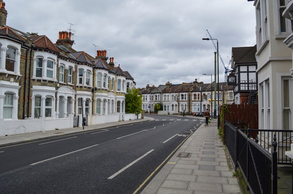 A quiet residential street in Bexleyheath with terraced Victorian-style houses featuring bay windows, brick and white-painted facades, and red-tiled roofs. The pavement is lined with a black metal fence and a row of concrete slabs, with a sidewalk on the right side. Overhead, a streetlight pole stands near the curb, and a construction crane is visible in the distance, indicating ongoing building works. The weather appears overcast with gray clouds, and no vehicles or pedestrians are present at the moment. This scene is suited for illustrating home relocation or moving services, such as those provided by Man and Van Bexleyheath, highlighting the typical environment for furniture transport and loading processes during a house removal.