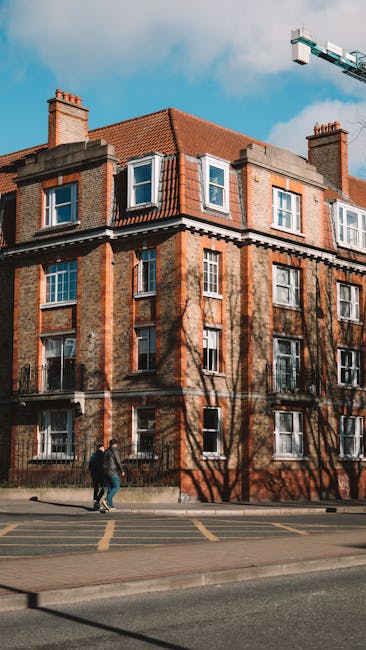 A multi-storey residential building with a red brick facade, white window frames, and a red tiled roof, situated on a street with a pavement. Shadows of leafless trees are cast onto the building's exterior. In front of the building, two individuals are walking on the pavement, carrying a cardboard box, possibly part of a house removal or relocation process. A construction crane is visible in the background against a partly cloudy sky. The scene depicts an environment suitable for house removals, with elements such as the building, sidewalk, and moving activity captured clearly, supporting the context of furniture transport and packing during home relocation, as managed by Man and Van Bexleyheath.