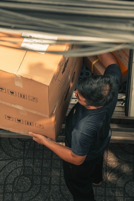 A man wearing a dark uniform is lifting a large cardboard box with printed symbols indicating fragile content and proper handling, inside the loading area of a van during a home relocation process. The box is wrapped with packing tape and positioned on the floor of the van, which has an open sliding door revealing the interior. The van's metal interior is visible at the top of the image, with the man using both hands to support the box, preparing for transportation. The scene is set on a patterned flooring surface beneath the open vehicle, with natural or ambient lighting illuminating the workspace. This image illustrates the logistics involved in professional furniture transport and packing during a residential move, as undertaken by Man and Van Bexleyheath, supporting efficient and careful house removals.