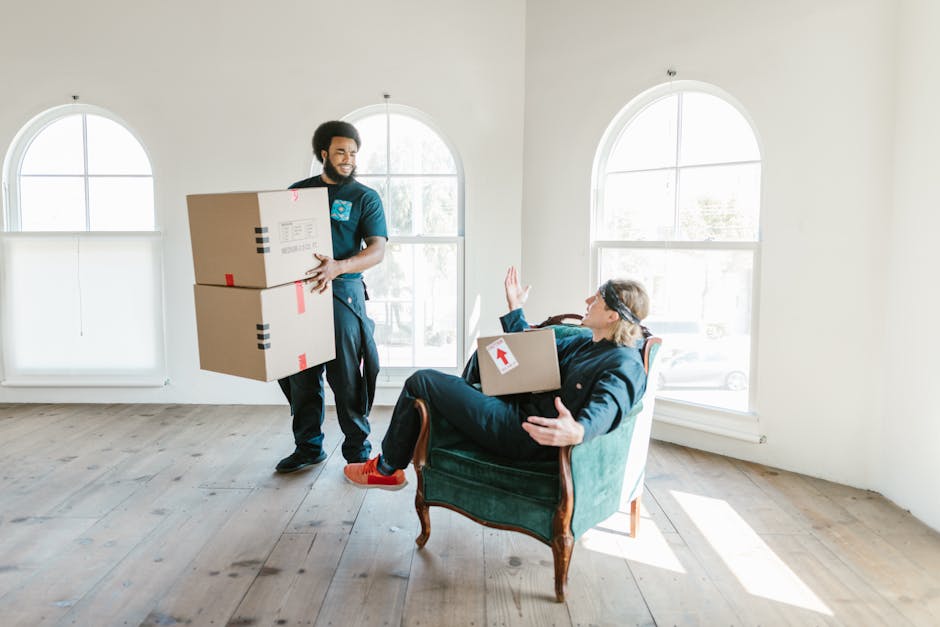 A man with dark curly hair and a beard, dressed in dark blue work clothes, is inside a bright, spacious room with large arched windows allowing natural light to illuminate the space. He is carrying two cardboard boxes stacked vertically, secured with red and black tape, and is walking across a wooden floor towards a man seated in an upholstered armchair. The seated man has short hair, glasses, and is wearing a dark jacket and red shoes; he is holding a box with a white arrow sticker, and has one hand raised in a gesture, possibly indicating a greeting or pause. The room has minimal furnishings, and the scene reflects a furniture transport or packing and moving process, illustrating professional removal services by Man and Van Bexleyheath during home relocation or office move.