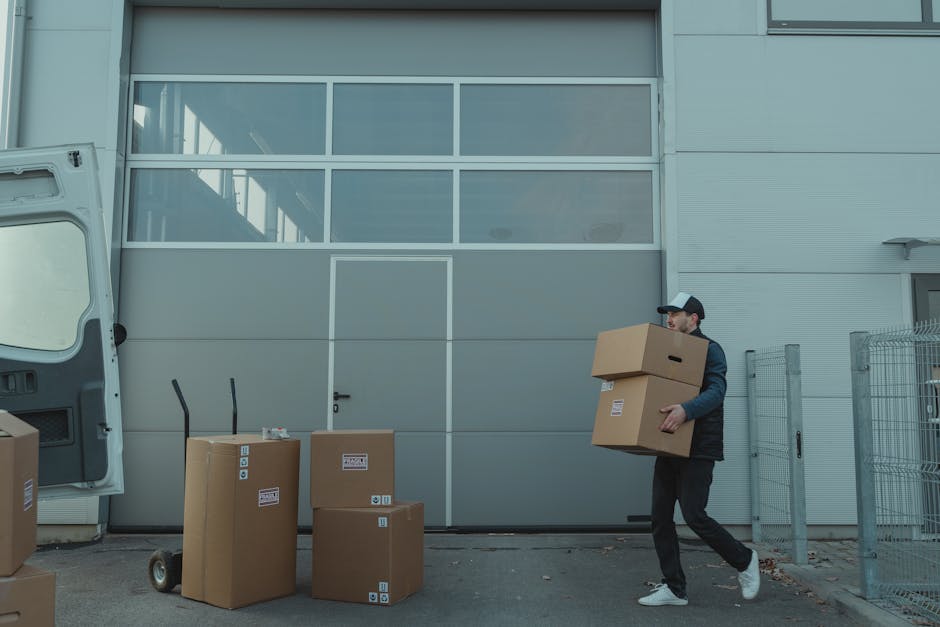 A man wearing a dark uniform is lifting a large cardboard box with printed symbols indicating fragile content and proper handling, inside the loading area of a van during a home relocation process. The box is wrapped with packing tape and positioned on the floor of the van, which has an open sliding door revealing the interior. The van's metal interior is visible at the top of the image, with the man using both hands to support the box, preparing for transportation. The scene is set on a patterned flooring surface beneath the open vehicle, with natural or ambient lighting illuminating the workspace. This image illustrates the logistics involved in professional furniture transport and packing during a residential move, as undertaken by Man and Van Bexleyheath, supporting efficient and careful house removals.