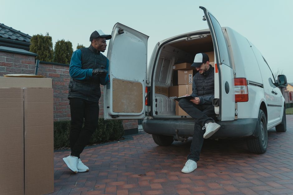 A young man with light brown hair and a beard, dressed in a blue hoodie over a white t-shirt and dark trousers, sitting on the edge of an open moving van parked on a driveway or street. He is holding 