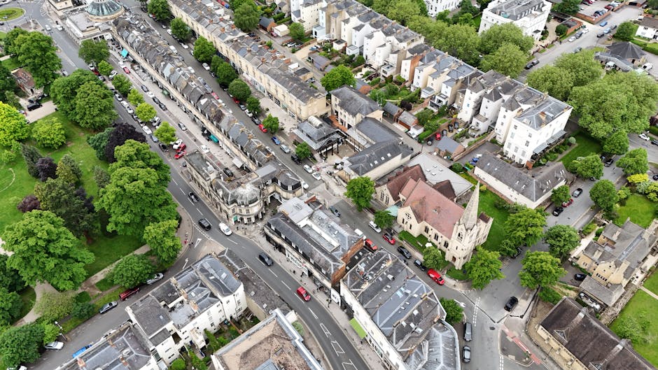 Aerial view of a residential neighbourhood with tightly packed terraced houses and detached buildings, intersected by narrow streets filled with parked cars and moving vehicles. Green trees line the sidewalks and fill the small parks between the houses, providing a lush, leafy environment. Near the centre, a church with a tall steeple is visible, surrounded by residential homes and some commercial buildings. The streets are busy with cars, some parked along the curbs and others in motion, reflecting typical urban traffic. The area appears well-maintained with tidy pavements, brick and stone facades, and neatly arranged greenery. This setting exemplifies a typical house relocation or furniture transport scene in Bexleyheath, where moving companies like Man and Van Bexleyheath may operate, managing loading and unloading processes within the neighborhood.