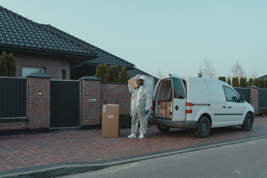 A mover dressed in full protective clothing, including a white disposable coverall, face mask, gloves, and shoe covers, stands outside a residential property during a home relocation process in Bexleyheath. The mover is carrying a cardboard box labeled for transport as part of packing and moving activities. Behind them, a metal cargo van with its side door open reveals filled cardboard boxes ready for loading onto the vehicle. The van is parked on a brick-paved driveway adjacent to a modern house with an asphalt tiled roof and a brick wall with a gate separating the property from the street. The scene takes place during daylight with overcast skies, and the surrounding environment includes neatly trimmed shrubs and some leafless trees in the background, emphasizing the logistical nature of furniture transport and packing during house removals, with the services of Man and Van Bexleyheath supporting efficient and secure home relocations.