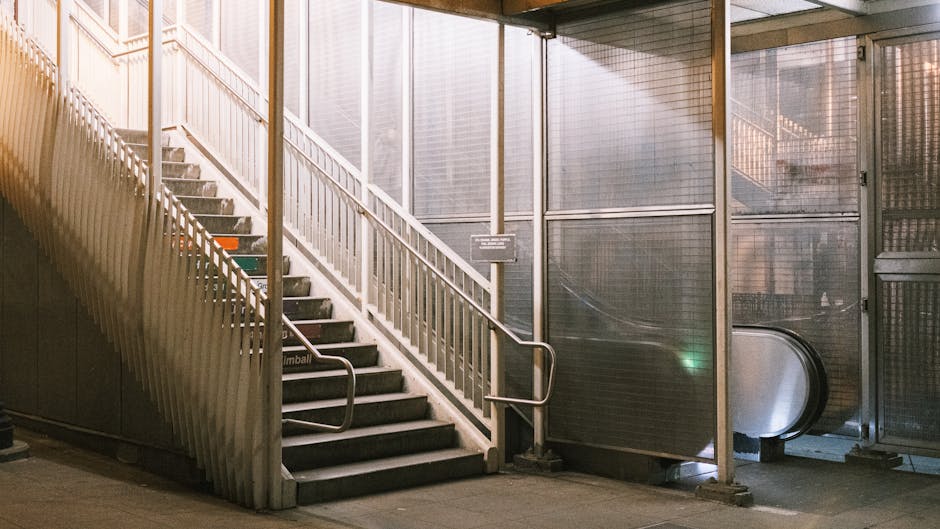 Image of a staircase with metal railings and concrete steps, situated inside a modern building near Bexleyheath station. The staircase curves upward to the left, with a white-painted handrail running along both sides. Adjacent to the staircase, small signs are mounted on the wall, possibly indicating directions or building information. To the right of the stairs, there is an escalator enclosed within metallic and glass structures, suggesting a transit or station environment. The lighting appears to be natural daylight filtering through nearby windows, illuminating the space with a neutral tone. This setting is relevant for house removals and furniture transport, as the staircase and escalator could be part of a home relocation process conducted by Man and Van Bexleyheath, providing insight into the logistics involved in moving household items through shared access points or public stations.