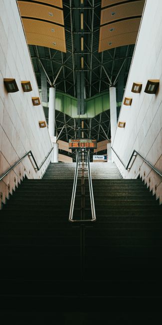 The image depicts a wide staircase inside a modern building with a high, dark-colored ceiling featuring a grid-like framework. The staircase has dark steps with a metal handrail running along the middle, leading upwards towards a landing. On either side of the staircase, there are white walls with rectangular, gold-colored wall sconces spaced evenly. At the top of the stairs, a sign with red text is partially visible, indicating directions or information. The interior lighting appears to be artificial, with a bright and neutral illumination enhancing the architectural details. This setting suggests an environment typical of public spaces such as a train station or transport hub, which aligns with the context of house relocations and furniture transport, as seen in services like those offered by Man and Van Bexleyheath.
