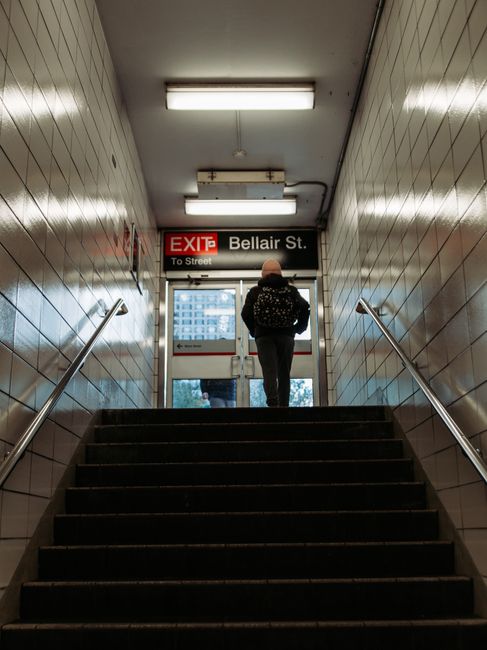 Image of a staircase with metal railings and concrete steps, situated inside a modern building near Bexleyheath station. The staircase curves upward to the left, with a white-painted handrail running along both sides. Adjacent to the staircase, small signs are mounted on the wall, possibly indicating directions or building information. To the right of the stairs, there is an escalator enclosed within metallic and glass structures, suggesting a transit or station environment. The lighting appears to be natural daylight filtering through nearby windows, illuminating the space with a neutral tone. This setting is relevant for house removals and furniture transport, as the staircase and escalator could be part of a home relocation process conducted by Man and Van Bexleyheath, providing insight into the logistics involved in moving household items through shared access points or public stations.