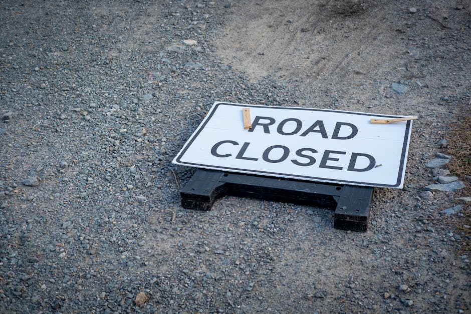 A black and white road closure sign with bold black text reading 'ROAD CLOSED' is placed on a gravel surface, secured with two wooden pegs at the top corners. The sign is positioned near the entrance of a property, indicating a temporary restriction on vehicle access which may impact home relocation or furniture transport activities. The surrounding area appears to be a construction or work zone with loose gravel and dirt visible in the background. The sign's placement suggests it is used to inform drivers and pedestrians about the road closure, which could be relevant during moving or packing and moving operations conducted by companies like Man and Van Bexleyheath. The lighting is natural, with no shadows or additional objects in view, emphasizing a straightforward message about the temporary traffic restriction at this location.