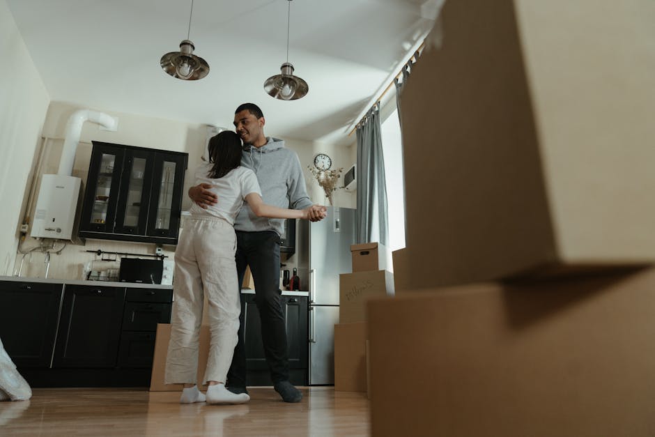 A man and a woman are inside a residential kitchen during a home relocation, with the man dressed in a grey hoodie and dark trousers, and the woman wearing a white top and light-colored pants. They are embracing each other, smiling, with the woman holding a small object in her hand. Several cardboard boxes are stacked on the wooden floor near the doorway, some labeled, indicating they are packed for moving. The kitchen features black and white cabinetry, a wall-mounted boiler, a fridge, and a wall clock with a decorative hanging plant nearby. Natural light streams in through a window with light blue curtains, illuminating the scene. The setting suggests the ongoing loading process of furniture and personal belongings, possibly facilitated by a professional removals service such as Man and Van Bexleyheath, involved in residential furniture transport and packing during house moves.