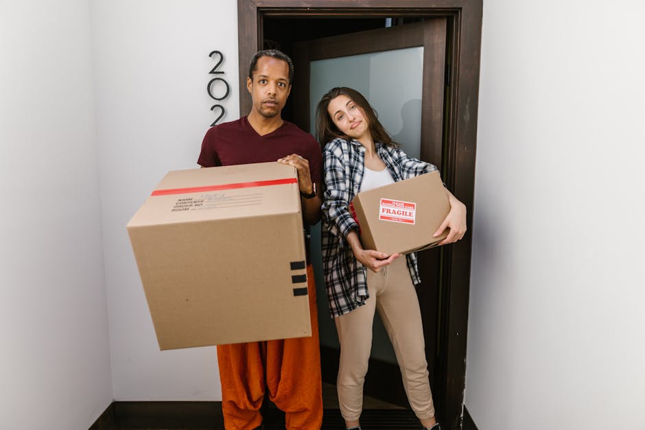 A man dressed in a white polo shirt and black cap, kneeling on the ground next to an open cardboard box, appears to be packing or writing on a piece of paper placed on the box. Several stacked cardboard boxes surround him, some sealed with packing tape, indicating preparation for a home relocation. The scene is set outside a property with an open garage or storage area, with natural daylight illuminating the workspace. In the background, a vehicle is partially visible, suggesting loading or unloading activity typical of furniture transport and packing processes during house removals. The man and van company, Man and Van Bexleyheath, is likely involved in this dismantling or packing stage of the moving logistics, demonstrating part of the loading process for a house removal service.